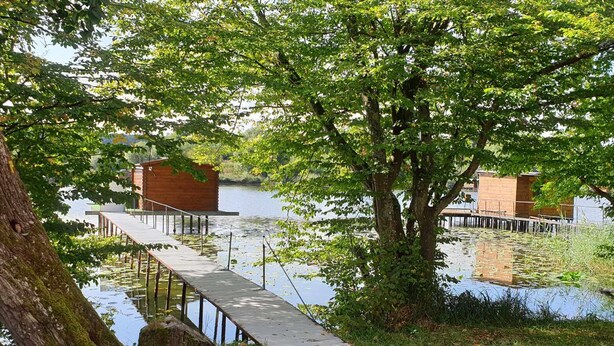 A wooden dock extending over a calm lake, surrounded by lush greenery and two wooden cabins.