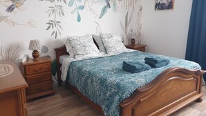 A neatly made bed with a floral bedspread, two matching pillows, a wooden nightstand with a lamp, and a wall with nature-themed wallpaper.
