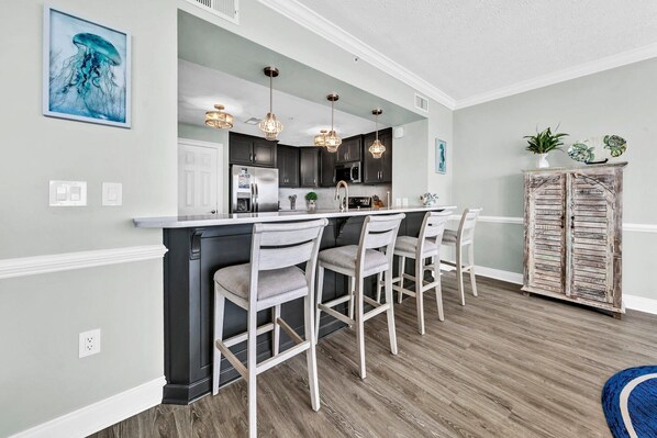 A modern kitchen with dark cabinets, a white island, and bar stools. There are pendant lights, a refrigerator, and a microwave.
