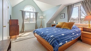 A bedroom with a sloped ceiling, a bed with a blue and white striped bedspread, a wooden nightstand, a lamp, and a window with curtains.