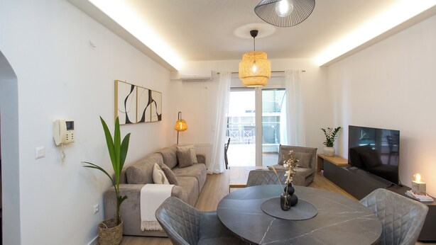 A modern living room with a round dining table, grey upholstered chairs, a grey sofa, a flat-screen TV, and a large potted plant. - K11 Apartment in Athens Downtown (Athina)