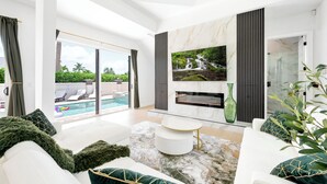 A modern living room with a pool view, a large TV, and a glass coffee table. - Naples Amore - Heated Pool/Spa, Pizza Oven, Playground, EV Charger, Grill, 5BR (Naples)