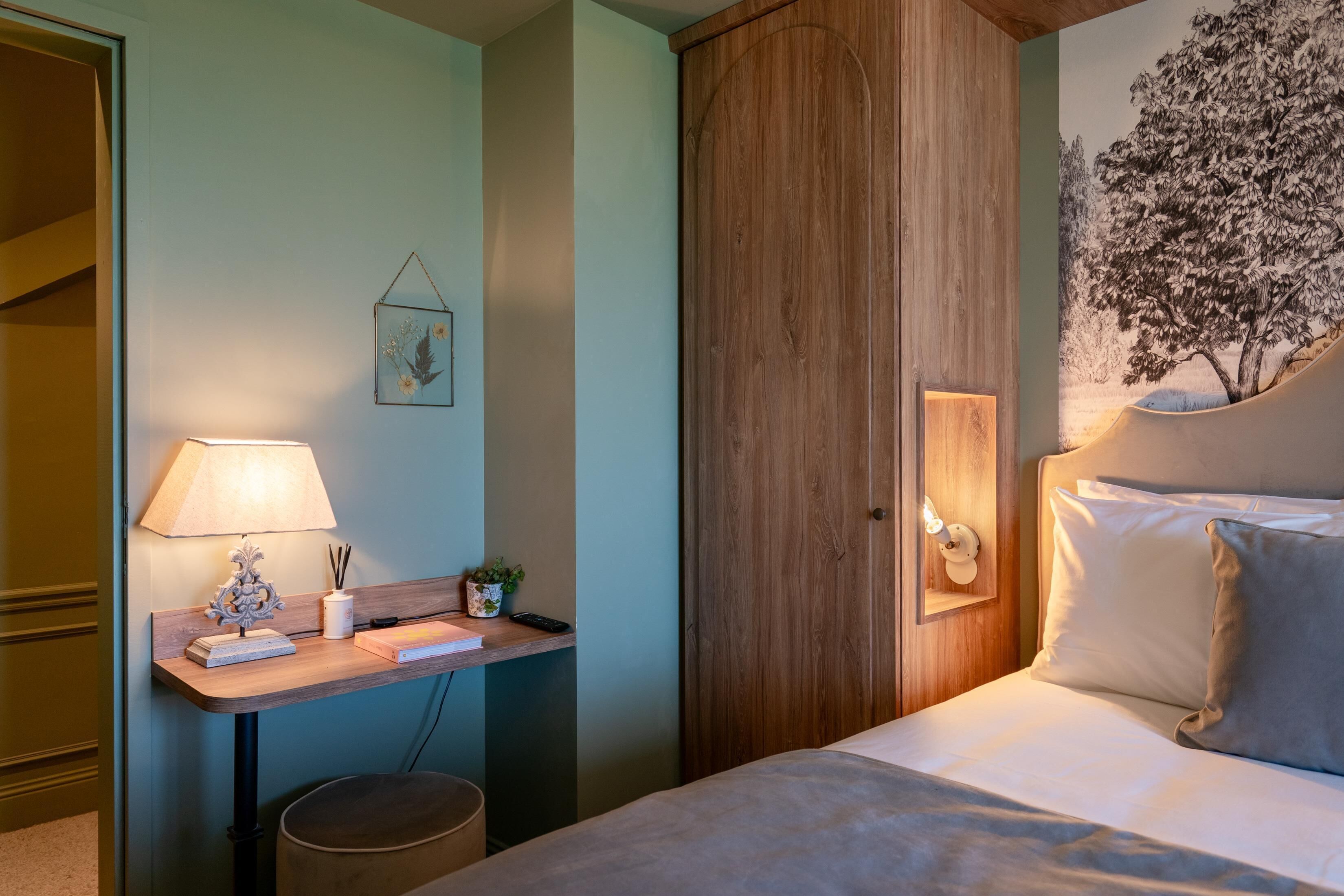 Un dormitorio con un escritorio de madera, una lámpara, una planta pequeña y una cama con almohadas.