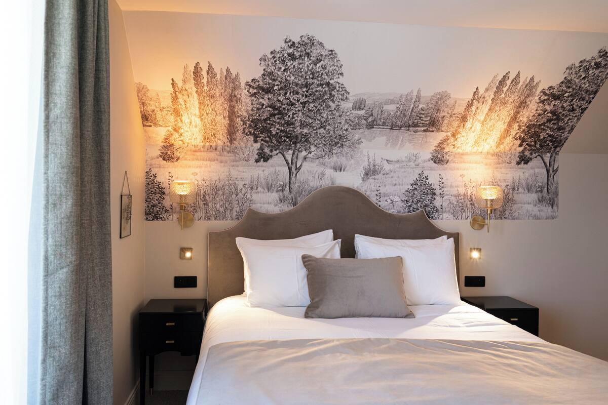 A neatly made bed with white linens and a single gray pillow, flanked by two bedside tables with lamps, in a room with a large landscape painting on the wall.