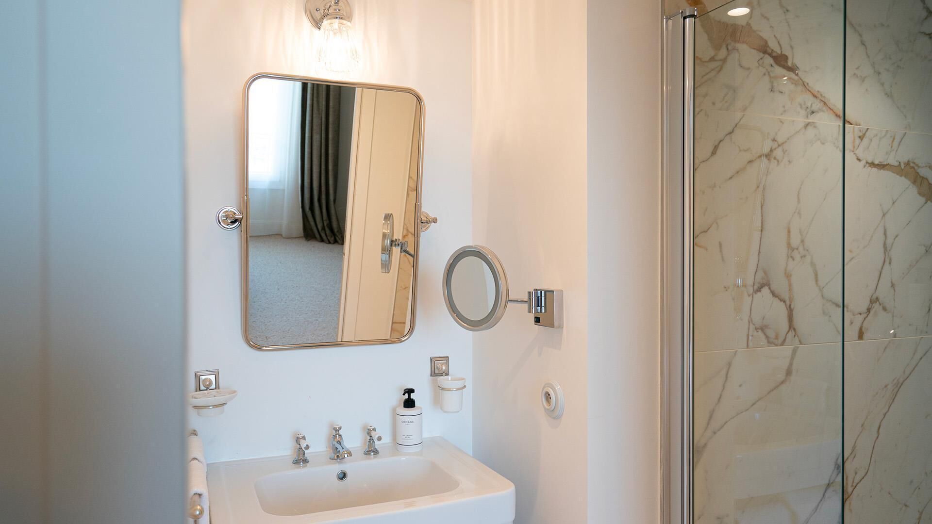 Une salle de bain avec un lavabo blanc, un miroir, une douche aux carreaux de marbre et un flacon de savon.