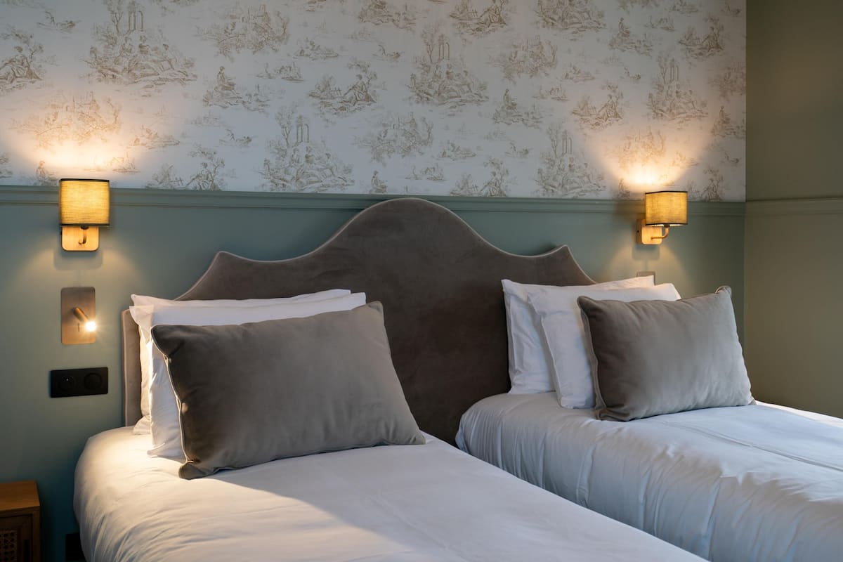 A hotel room with two beds, a headboard, wall sconces, and a patterned wallpaper.