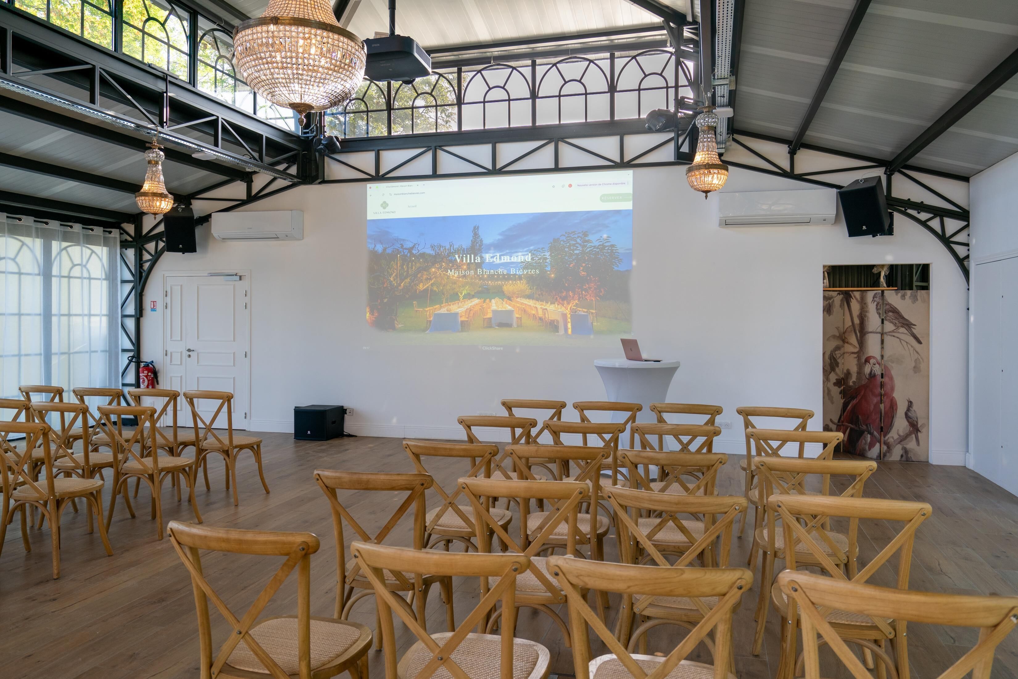 Een ruime kamer met houten stoelen, klaar voor een evenement, een projectiescherm waarop een landschap wordt vertoond, en een groot schilderij aan de muur.