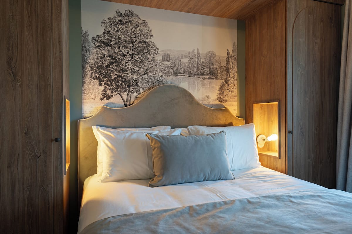 A bed with white linens and pillows, a headboard with a light fixture, and a mural of a winter landscape on the wall behind.