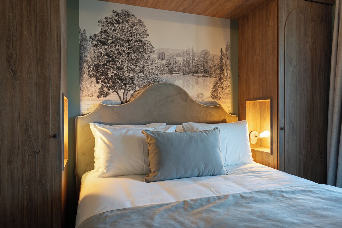 Un lit avec du linge de lit blanc et des oreillers, une tête de lit dotée d’un luminaire et une fresque représentant un paysage hivernal sur le mur derrière.