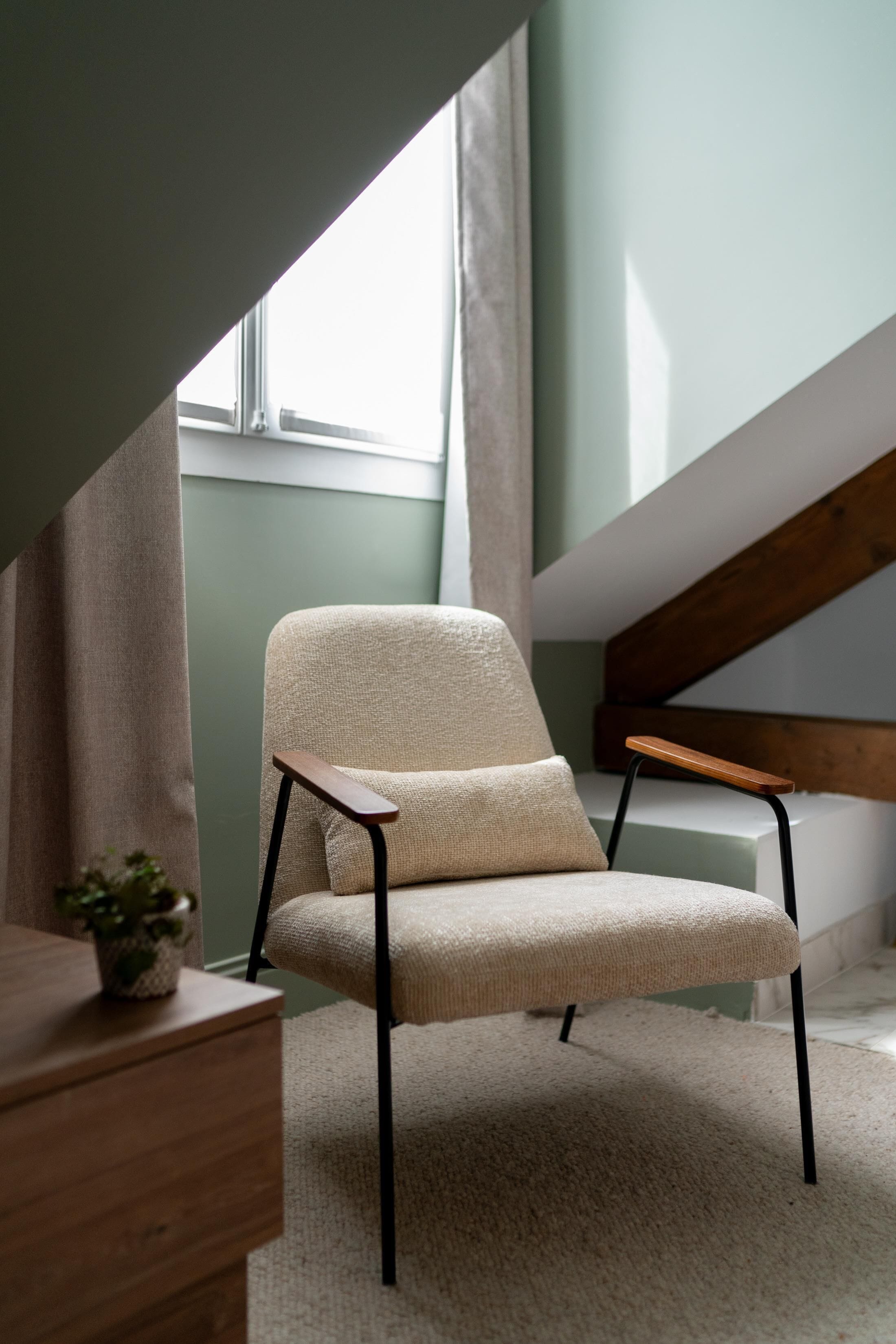 A modern chair with a wooden armrest in a room with a sloped ceiling and a window.