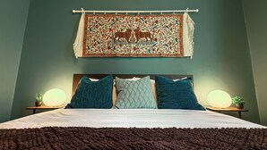 Een bed met een donkerbruine deken, twee blauwe kussens en een decoratief wandkleed met een tijger en bloemenmotief erboven.