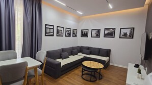 Ein modernes Wohnzimmer mit Ecksofa, einem kleinen runden Tisch und einem Esstisch mit Stühlen. An der Wand befinden sich gerahmte Bilder und ein Flachbildfernseher.