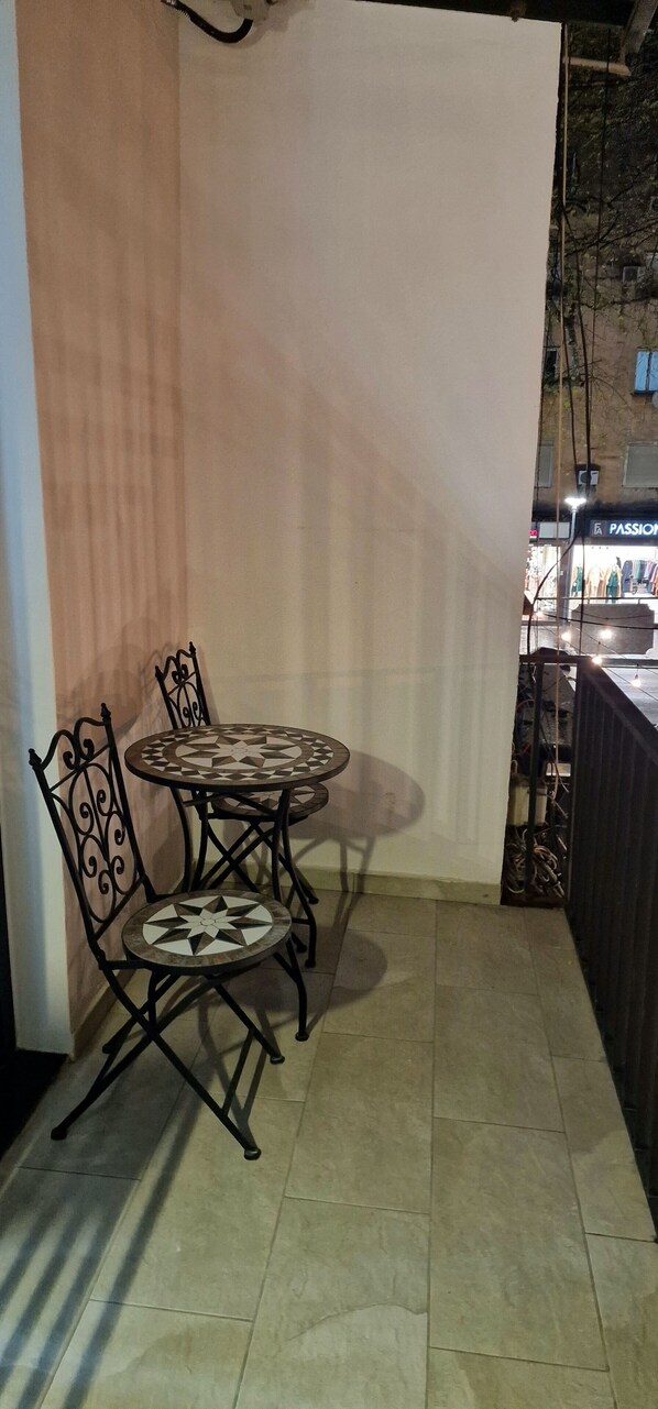 A small patio area with a round table and two chairs, a decorative metal chair, and a view of a street with shops. - Arkle Apartment 6 (Tirana)