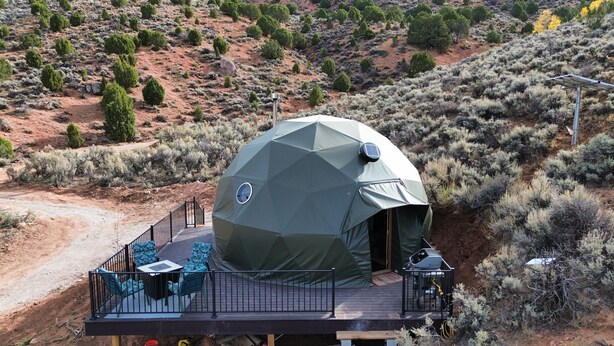 A geodesic dome tent with a deck, furnished with a table and chairs, surrounded by desert vegetation.