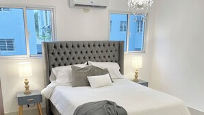 Un dormitorio ordenado con una cama grande, dos mesitas de noche, una araña y vistas a través de las ventanas.