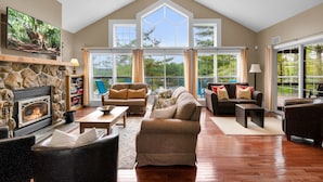 A spacious living room with a stone fireplace, comfortable seating, large windows, and a view of greenery.