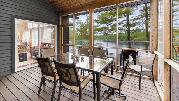 A covered outdoor seating area with a glass table and chairs, overlooking a body of water and trees.