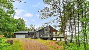 A two-story cabin with a covered deck and a garage, surrounded by a forest and a lake.