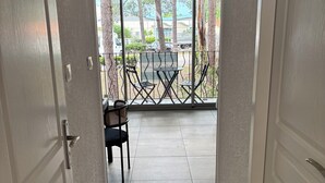 Un couloir mène à un balcon où se trouvent une table et des chaises. De là, on peut admirer la vue sur les arbres et les voitures garées, ainsi que sur un bâtiment situé au loin.