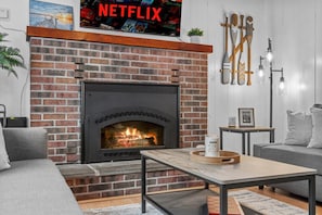 A living room with a brick fireplace, a grey sofa, a coffee table, and a wall-mounted TV displaying the Netflix logo.