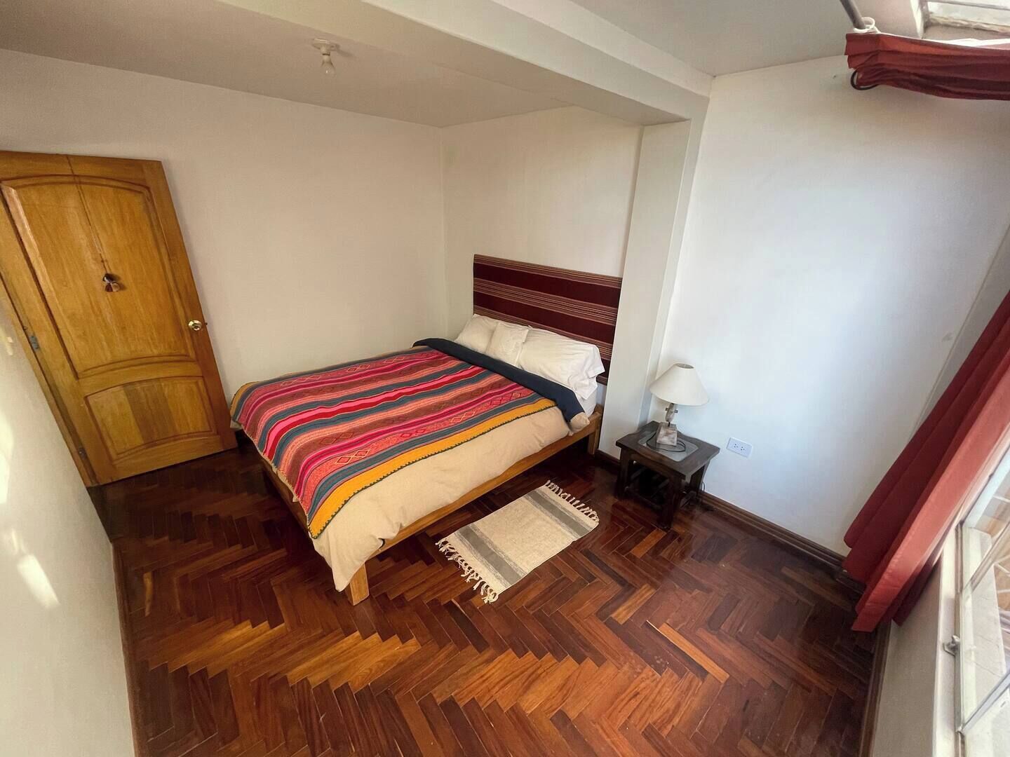 Une chambre avec un plancher en bois, un lit recouvert d’une couverture à rayures, une table de chevet avec une lampe et une porte en bois.