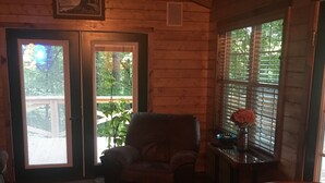 A cozy cabin interior with a leather armchair, a wooden table, and a view of the outdoors through a door and window. - Cozy 2-bedroom cabin in charming Whittier with WiFi, AC (Whittier)