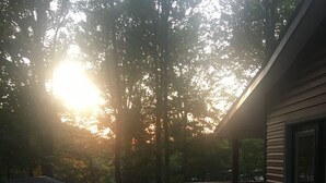 A wooden deck with a railing, leading to a covered area with a view of trees and the sun setting in the background. - Cozy 2-bedroom cabin in charming Whittier with WiFi, AC (Whittier)
