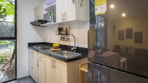 A modern kitchen with a black countertop, stainless steel sink, and wooden cabinets. There is a refrigerator, a small table with a checkered tablecloth, and a window with a view of greenery.