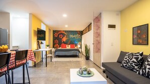 A modern living room with a sofa, a dining table with chairs, a bed with a colorful mural, and a kitchen area.