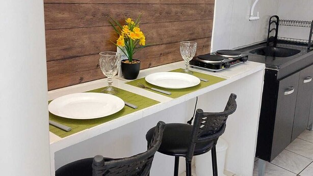 A compact kitchen with a wooden accent wall, a small dining area with two black bar stools, a white countertop, and a sink. - APARTMENT ACONCHEGO, NEXT TO NORTH SHOPPING AND DOWNTOWN MARACANAU (Jereissati II Maracanaú)