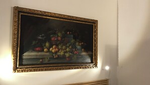 A bedroom with a bed, white pillows, a nightstand, a small round table, and a framed painting of fruits and vegetables on the wall.