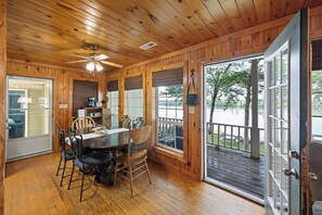 Un interior de madera con comedor, ventilador de techo y amplias ventanas con vistas a un lago.