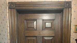 Una puerta de madera oscura con el número '5' y una cerradura, flanqueada por molduras decorativas y una pared con papel tapiz floral.