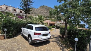 A white SUV parked on a stone pathway in a residential area with houses, trees, and a mountainous backdrop. - Lighthouse Villa (Santa Flavia)