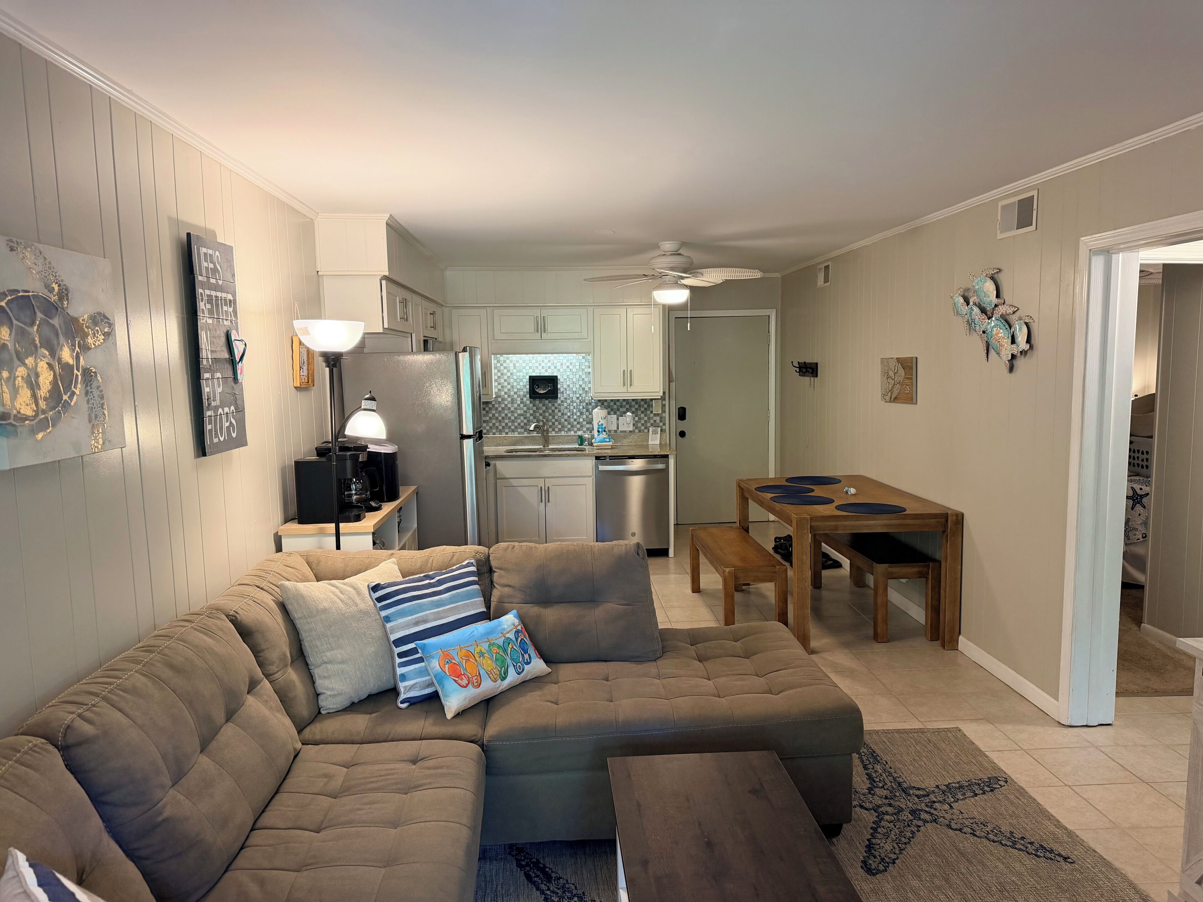 A living room with a sectional sofa, a dining table with four chairs, a kitchen in the background, and a ceiling fan.