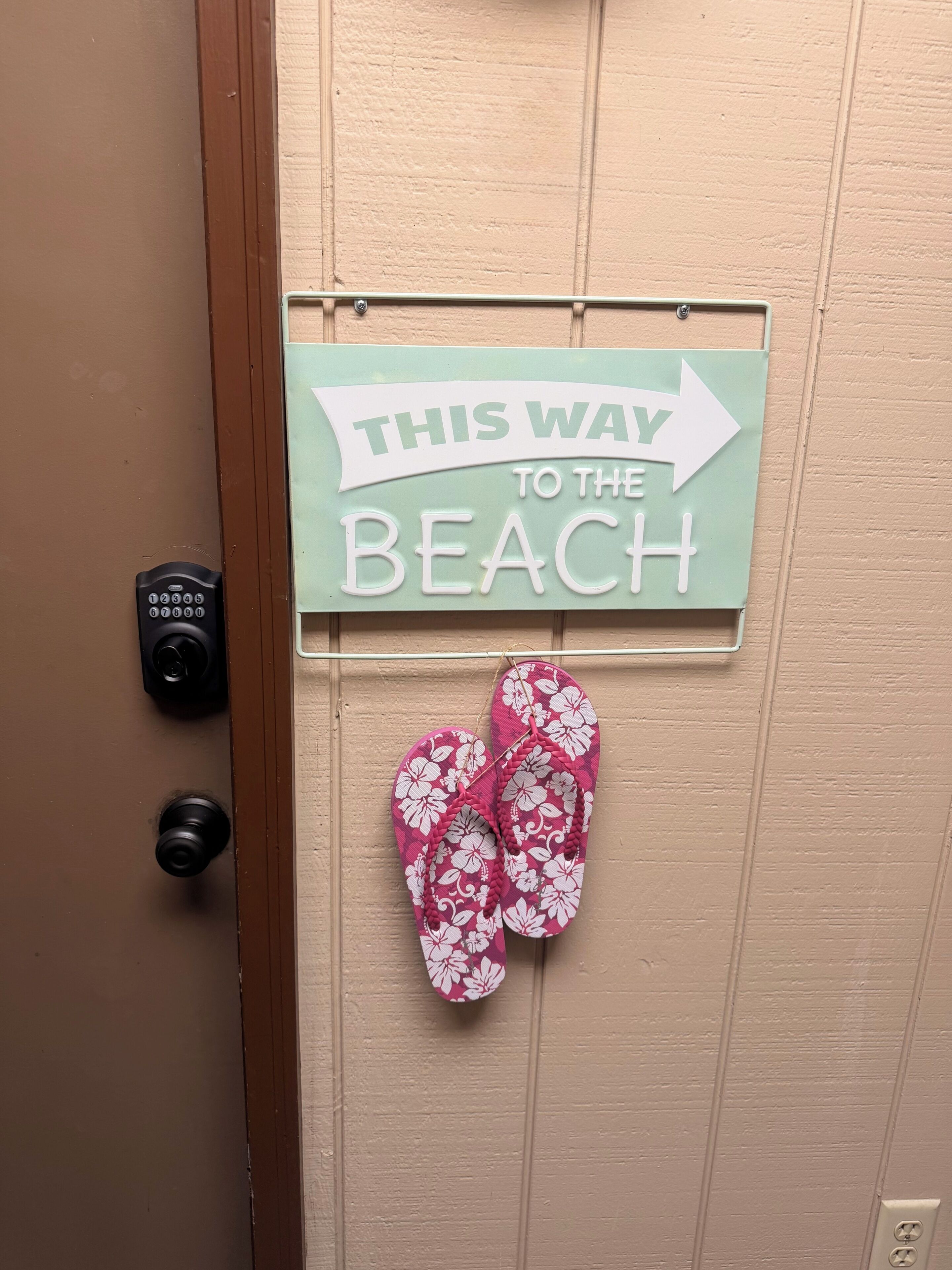 Une porte sur laquelle est affichée une pancarte indiquant la direction de la plage, et une paire de tongs suspendues à la porte.