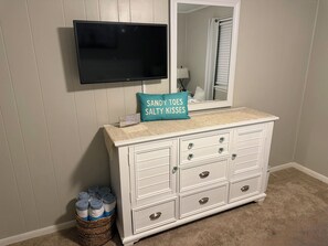 A white dresser with drawers, a flat-screen TV mounted above it, a teal pillow with text, and a wicker basket with folded towels. - Flip-Flop Getaways-Steps to Beach-Pool-King Bed (Hilton Head Island)
