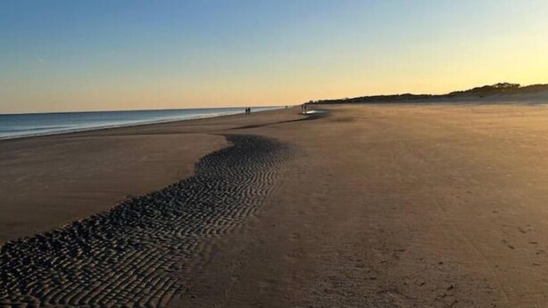 A beach with a long stretch of pebbles leading to the water, a clear sky, and a distant shoreline.