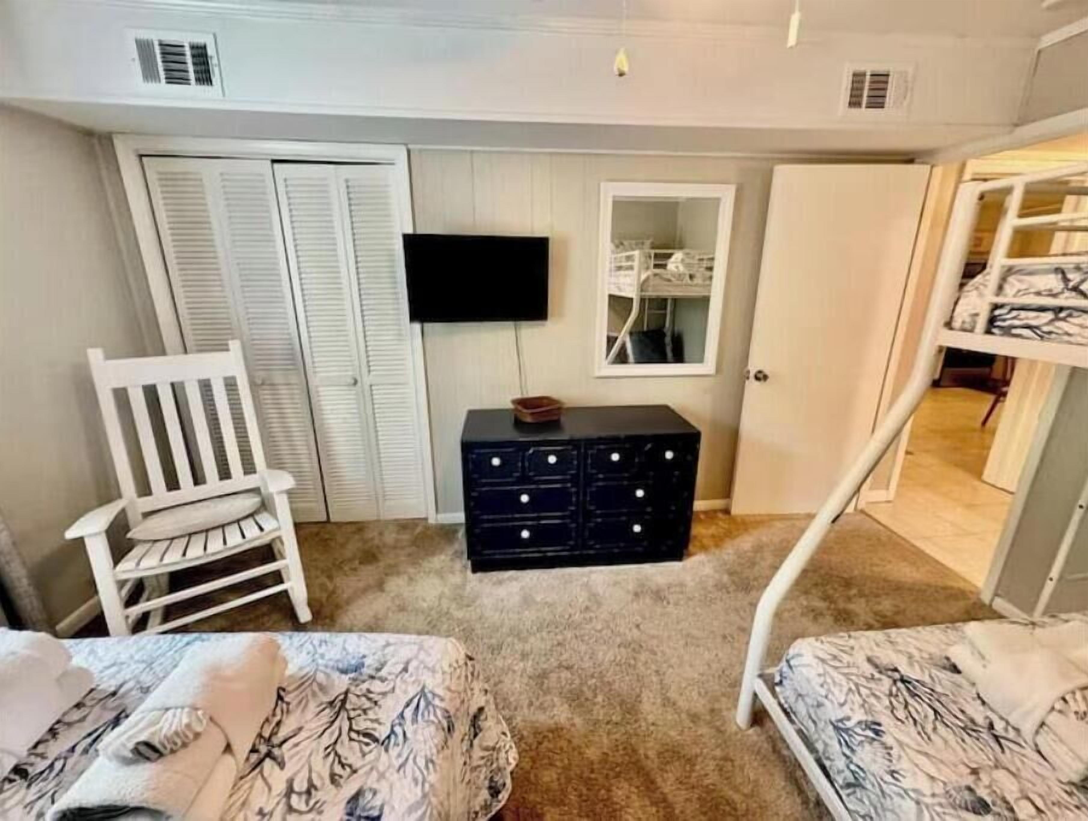 Een slaapkamer met twee bedden, een witte stoel, een dressoir, een televisie en een kast.