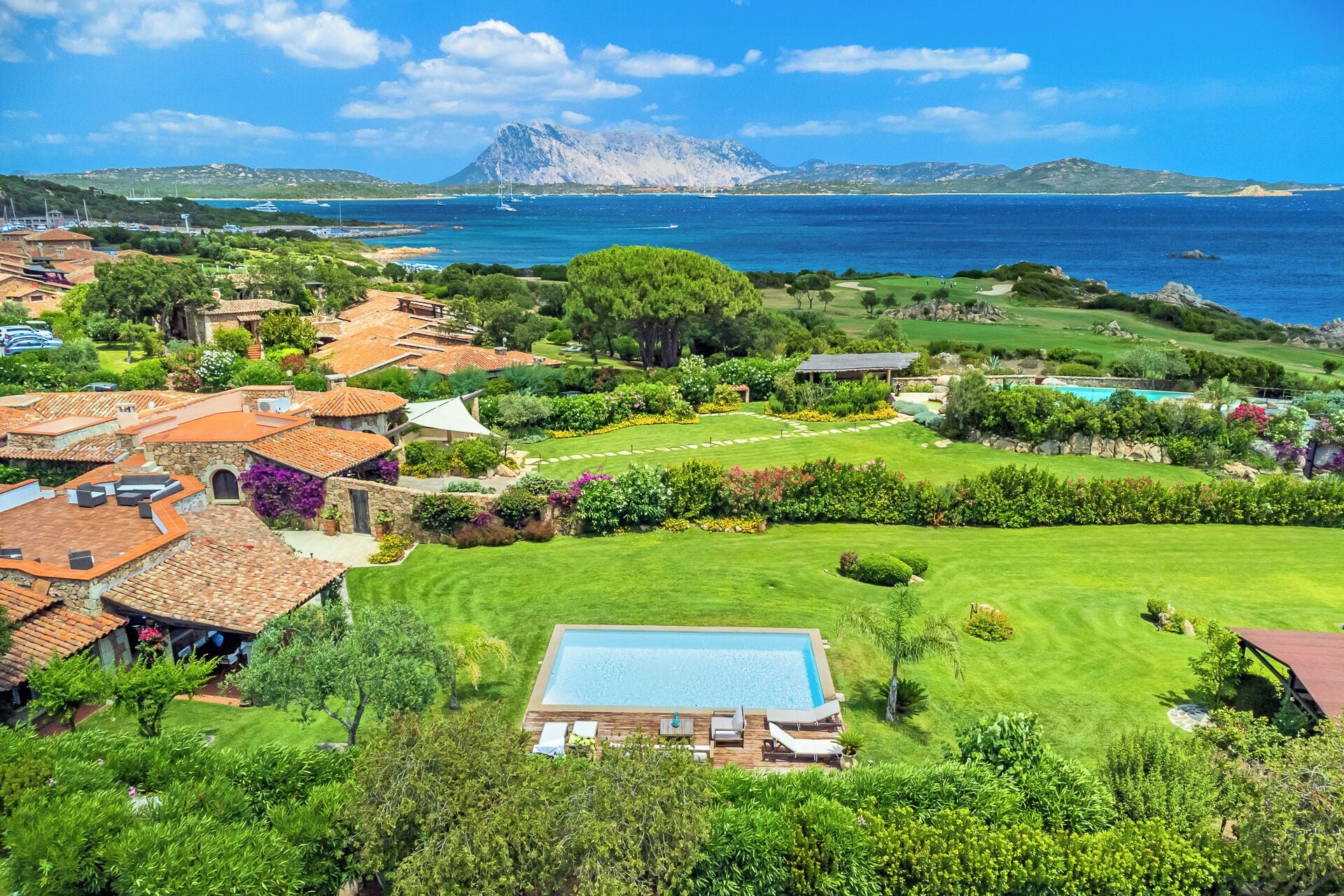 A coastal property with a swimming pool, lush gardens, and a view of the sea and mountains.