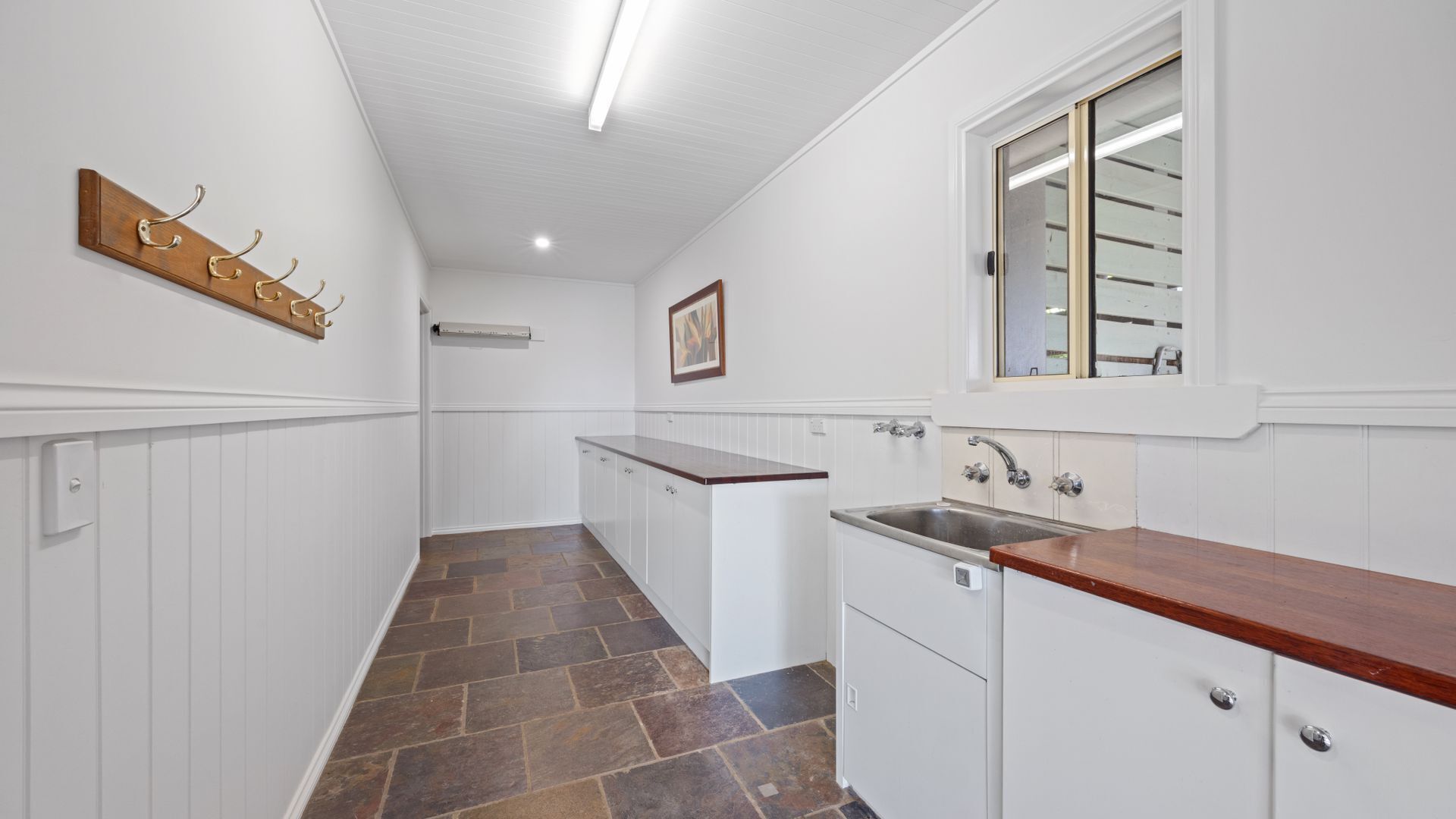 Un pasillo estrecho con suelo de madera, paredes blancas, un perchero de madera, un cuadro enmarco, una ventana con espejo y una zona de cocina con fregadero y armarios.