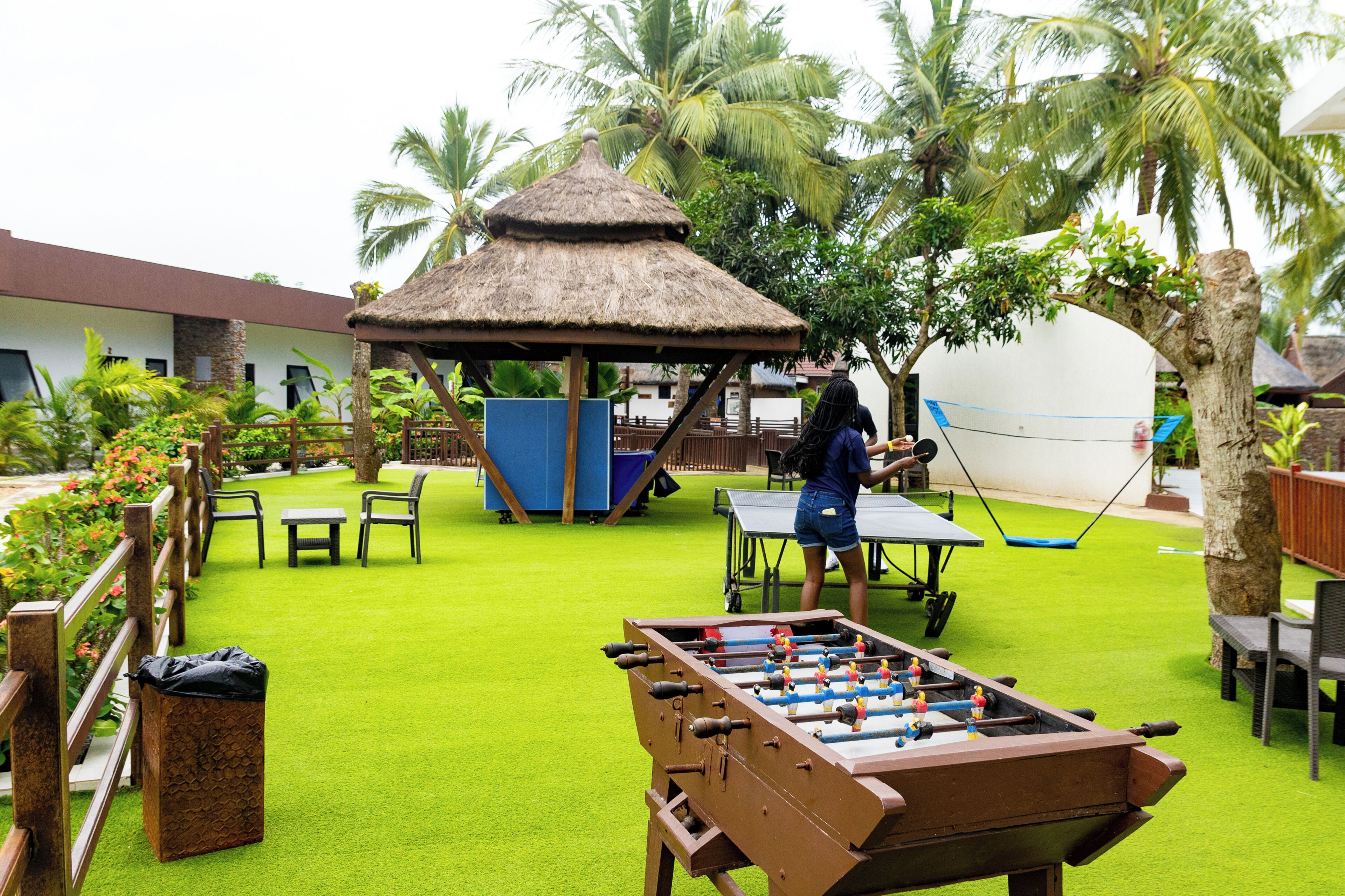 An outdoor recreational area with a foosball table, a thatched-roof gazebo, and a table tennis table.
