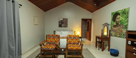 A room with a bed, two armchairs, a table, a ceiling fan, and a painting on the wall.