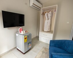 A room with a flat-screen TV mounted on the wall, a mini-fridge with a sticker, a blue sofa, and a walk-in closet with hanging robes.