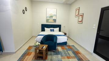 A hotel room with a bed, a chair, a small table with a vase and remote controls, and artwork on the walls.