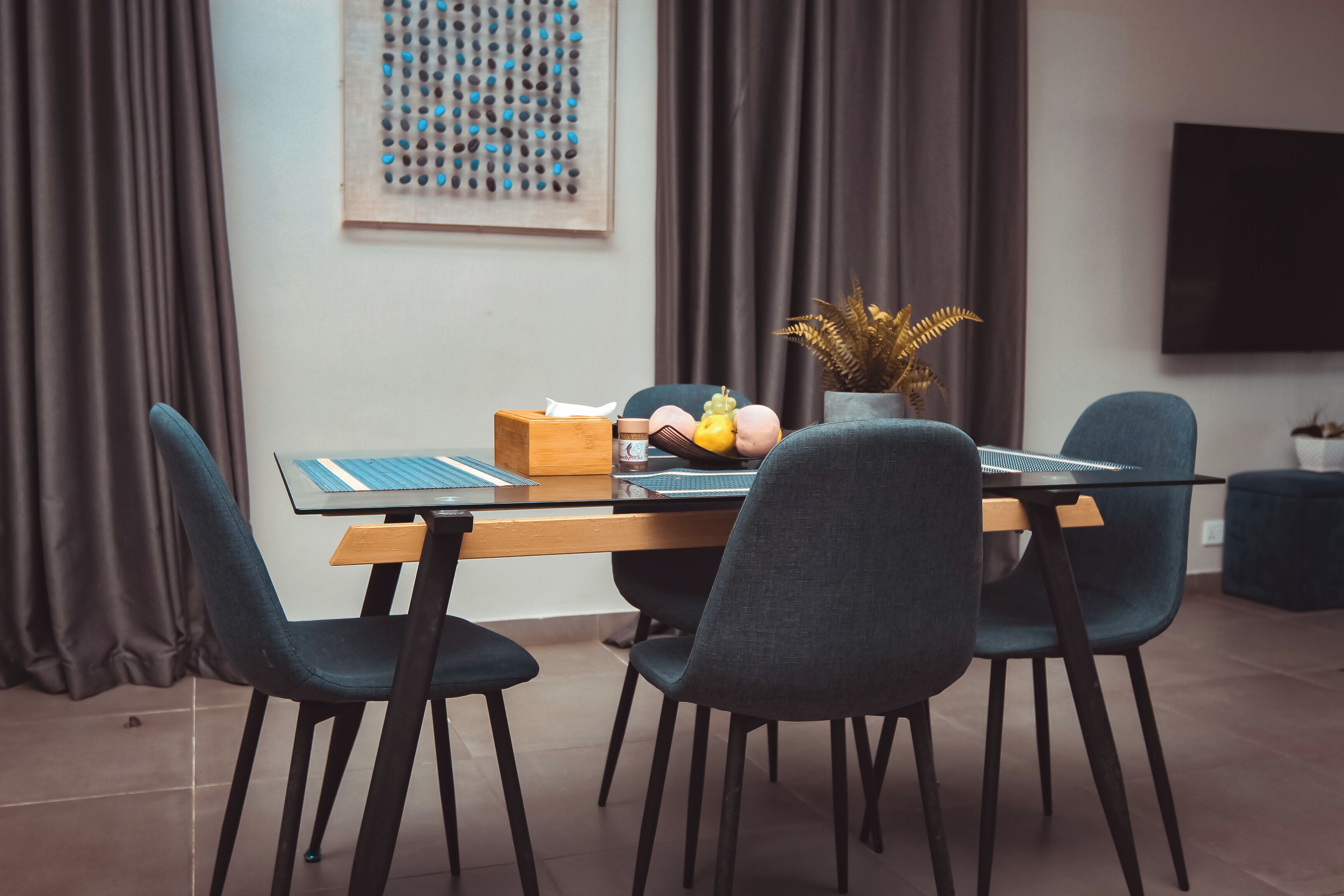 A modern dining area with a glass table, dark chairs, a potted plant, and a flat-screen TV mounted on the wall.