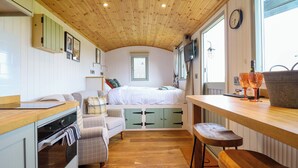 A compact, well-lit room with a wooden ceiling, a bed with white bedding, a kitchenette with an oven, a dining area with a table and chairs, and a window with curtains. - Charollais (Oswestry)