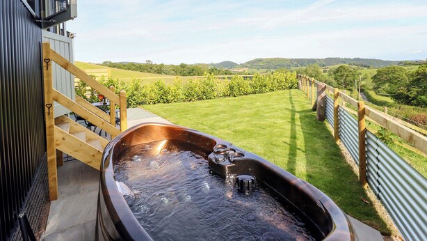 A hot tub with a wooden staircase leading up to it, surrounded by a metal fence and a view of a green landscape. - Charollais (Oswestry)