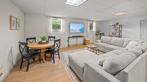 A modern living room with a dining area, a grey sofa, a flat-screen TV, and a bookshelf.