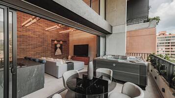 A modern balcony with a glass door leading to a living area, a hot tub, and a seating area with a table and chairs.